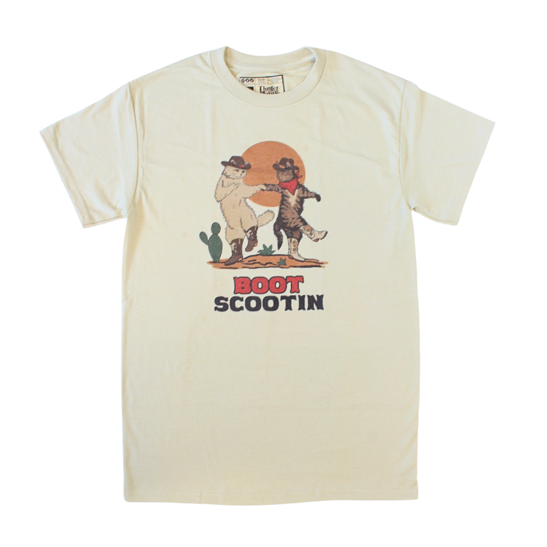 Two cats in cowboy hats and boots dancing in the desert. Natural tan shirt says, "Boot Scootin" country western font. 