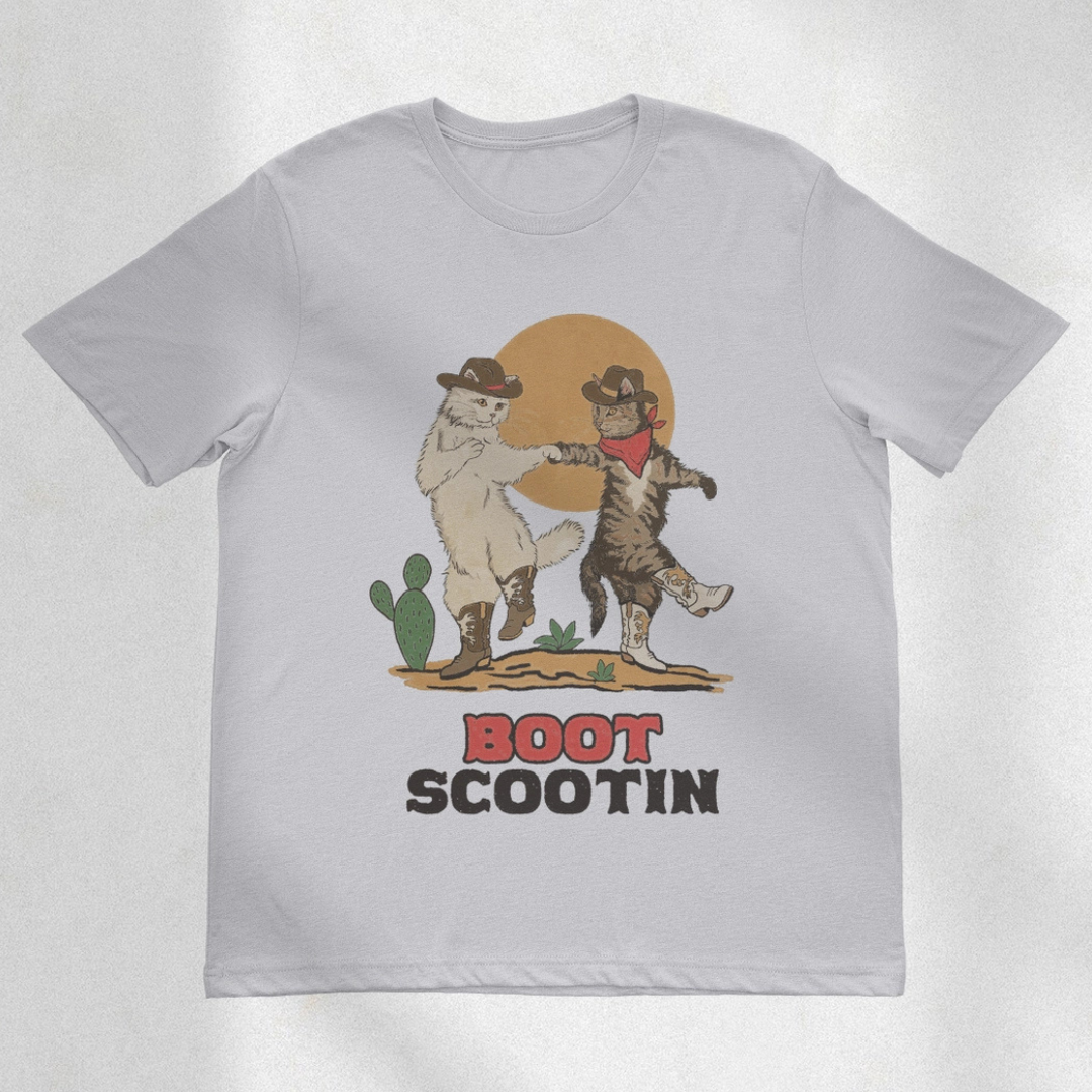 Two cats in cowboy hats and boots dancing in the desert. Natural tan shirt says, "Boot Scootin" country western font. 