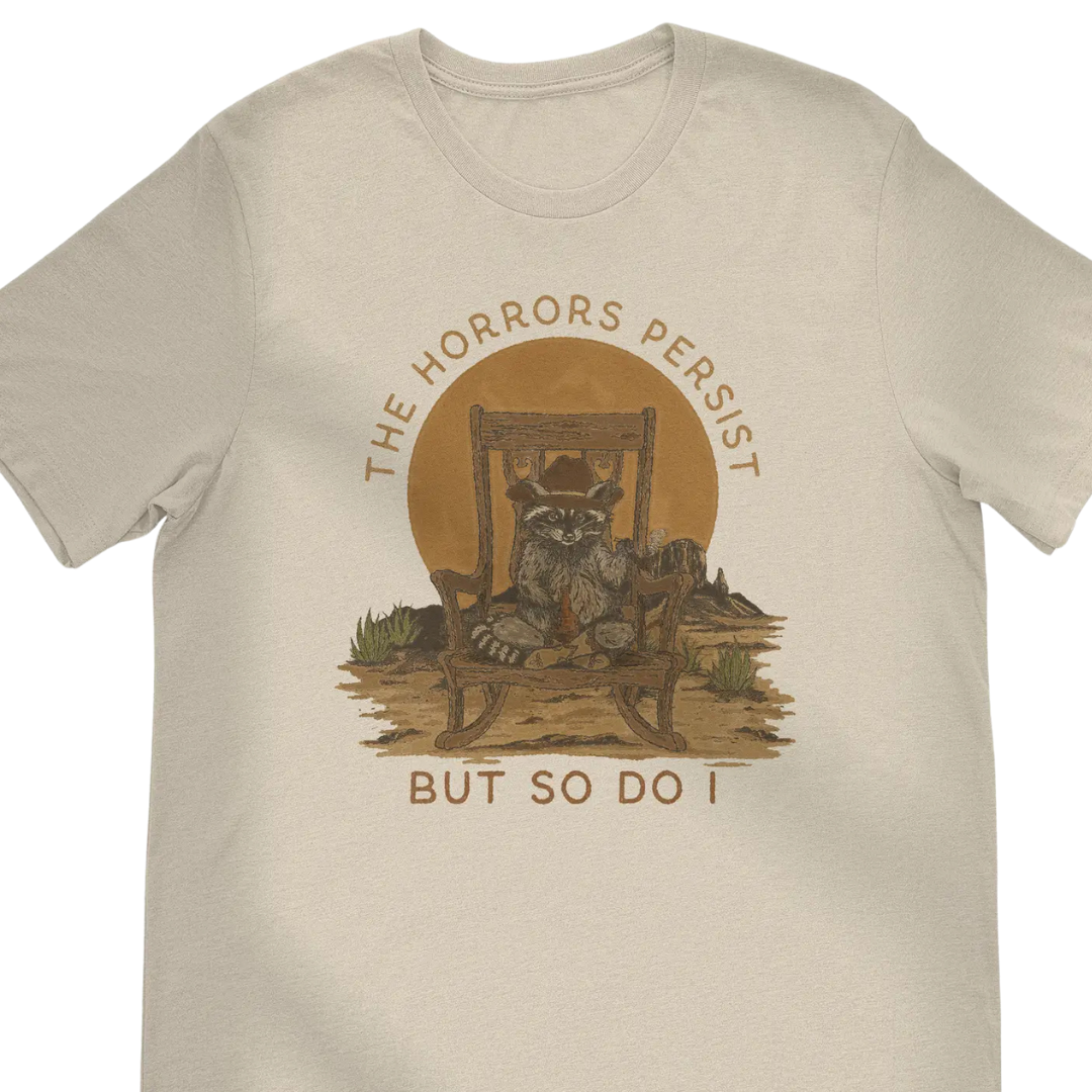 Tan shirt with Racoon cowboy in a rocking chair "The Horrors Persist but so do I".