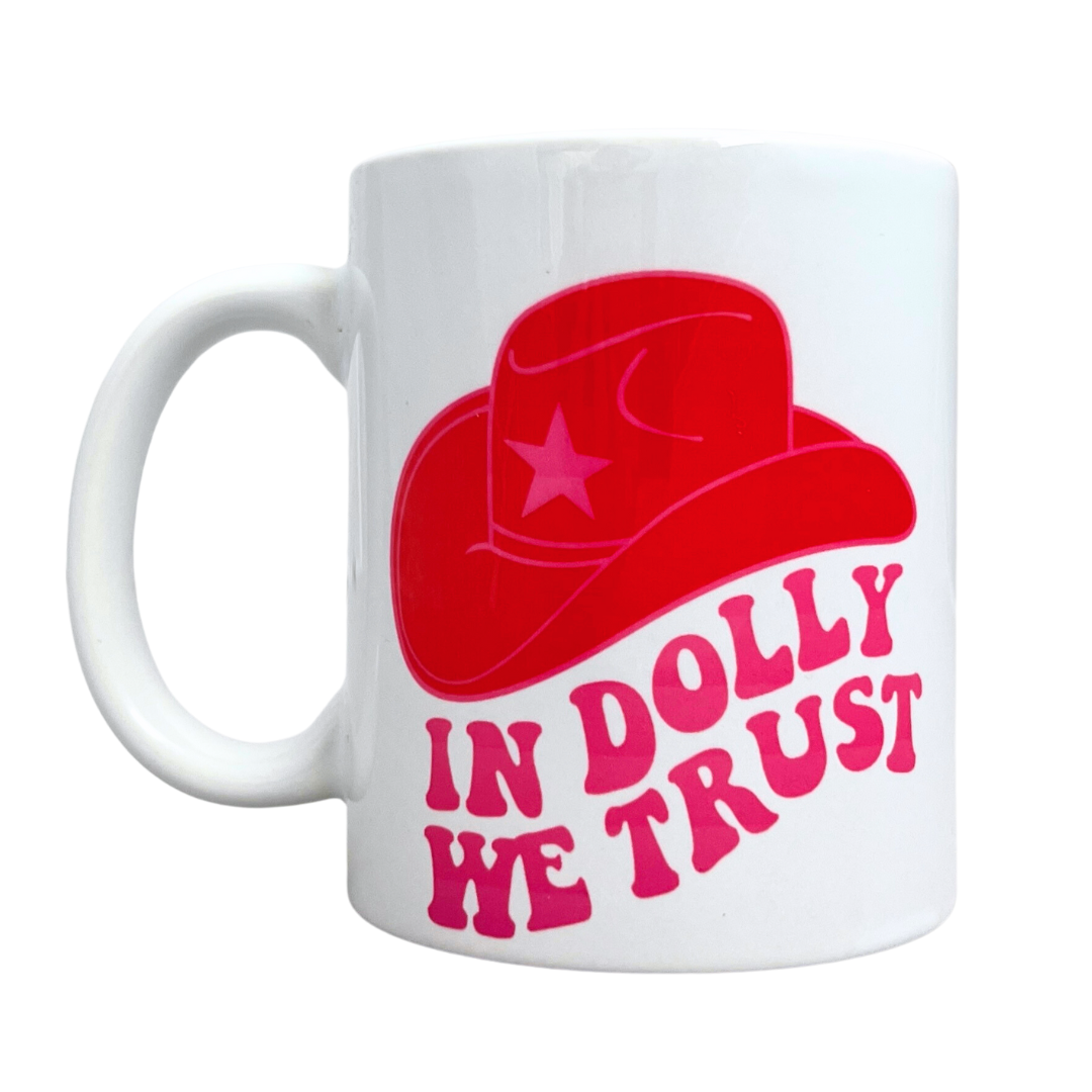White mug with "In Dolly We Trust" in a groovy pink font. Features a red cowboy hat as well.